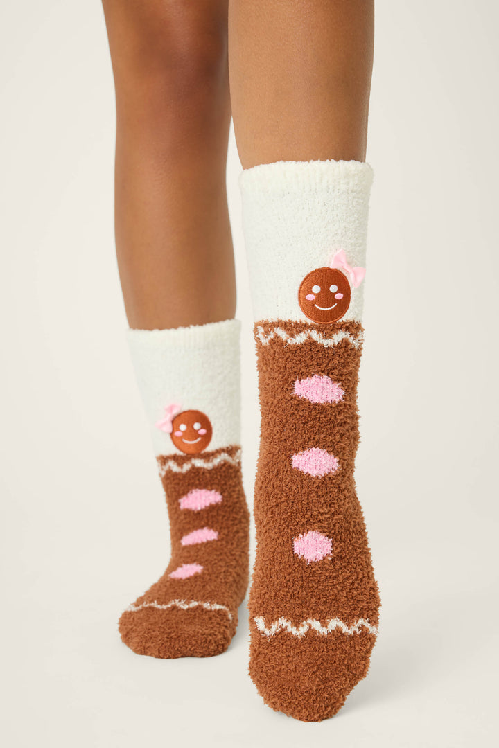 Someone wears P.J. Salvage GINGERBREAD MAN SOCKS SKI JAMMIE CLASSICS—fuzzy white and brown socks with pink dots, a gingerbread cookie face, and a pink bow—against a plain, light background.