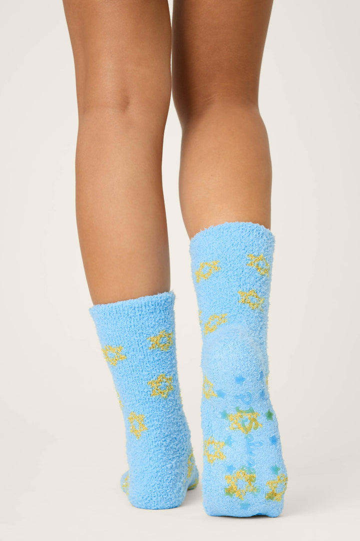 A person stands with their back to the camera, showing only their legs and feet in P.J. Salvage STARS SOCKS BOYFRIEND BOXER SHORT GIFT SETS light blue fuzzy socks with yellow and green sun shapes, against a plain light background.