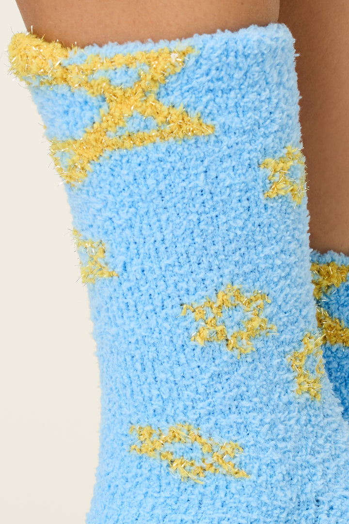 A close-up of a leg wearing P.J. Salvage Star Of David Print Crew Socks with Grippers, featuring a light blue fuzzy design with yellow textured patterns. The background is plain and light-colored.