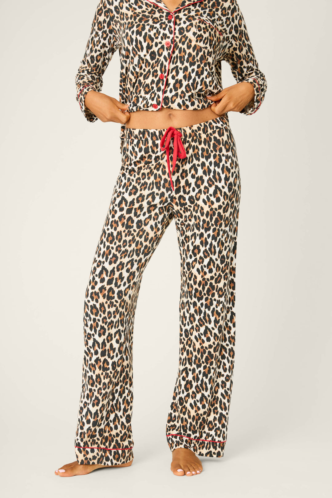 A person stands barefoot wearing P.J. Salvage Holiday Dreams Leopard Print Open Leg Pants, styled with a matching long-sleeve top featuring red trim and buttons. The person's head is not shown in the image.