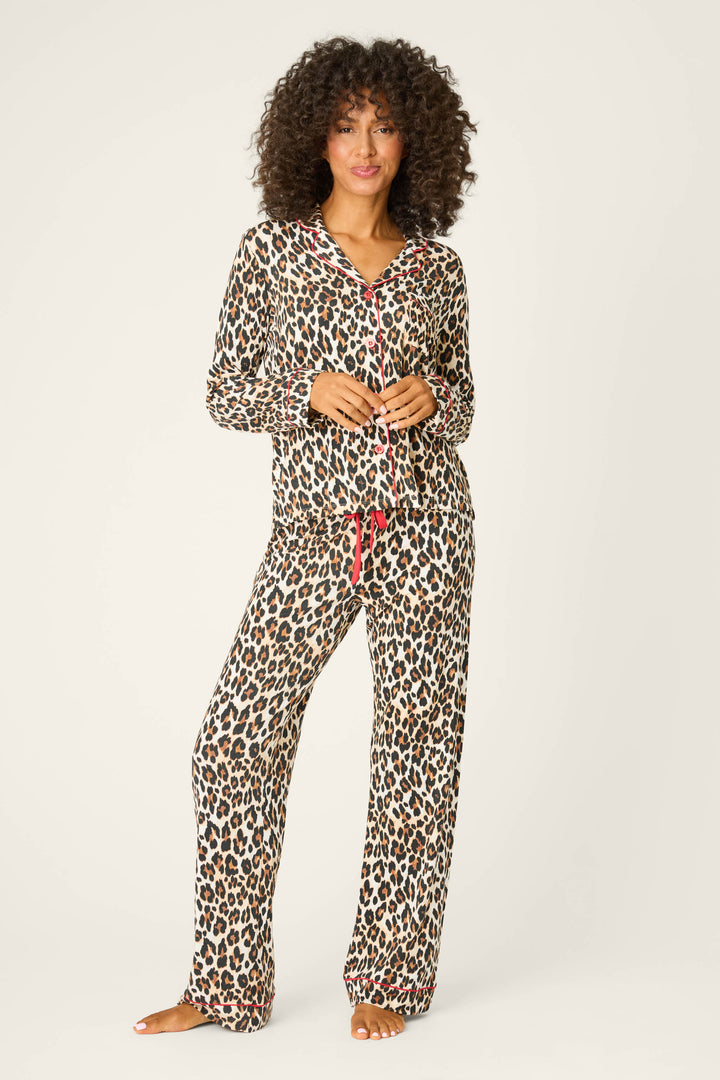 A person stands barefoot against a plain background, wearing the P.J. Salvage LEOPARD PJ SET HOLIDAY DREA— a matching leopard print pajama set with pink trim, long-sleeved top, and loose-fitting pants.