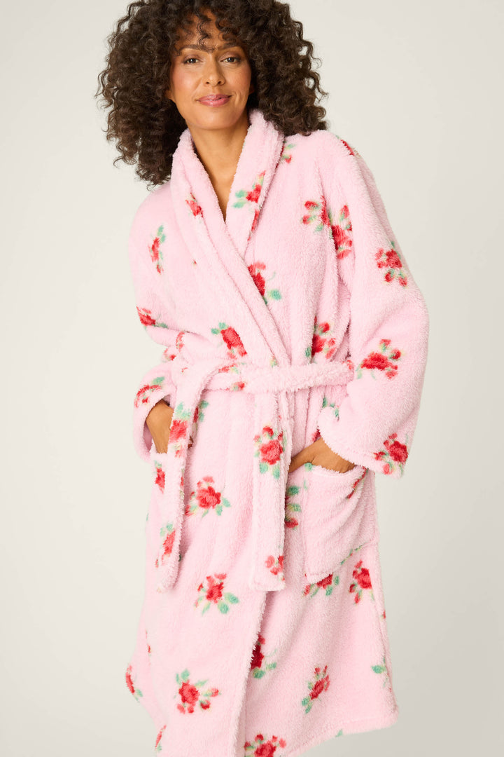 A person smiles at the camera wearing the P.J. Salvage Rose Print Plush Robe, featuring light pink fabric with red and green floral patterns, large front pockets, a tied waist belt, and one hand tucked into a pocket.