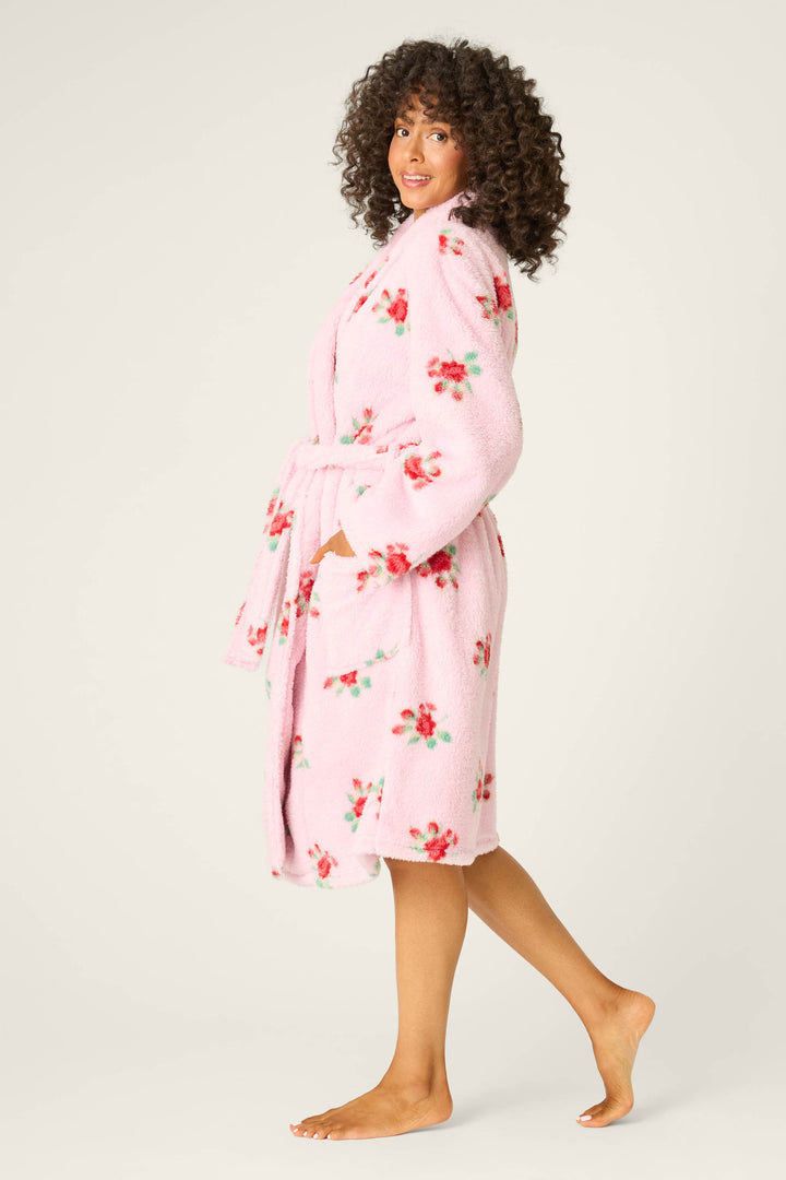 A person with curly hair models the P.J. Salvage Rose Print Plush Robe—pink, floral, knee-length with a belt and pockets—standing barefoot and sideways against a plain light background.