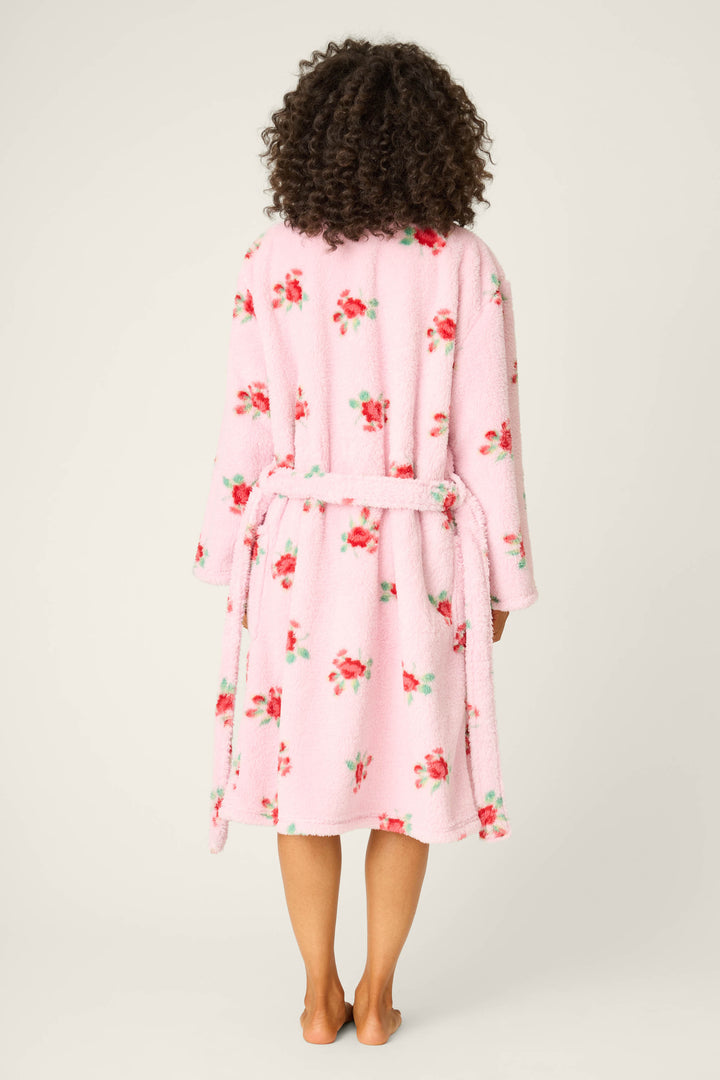 A person with curly hair stands barefoot, facing away, wearing the P.J. Salvage Rose Print Plush Robe—a pink floral robe with a tie belt—against a plain, light-colored background.