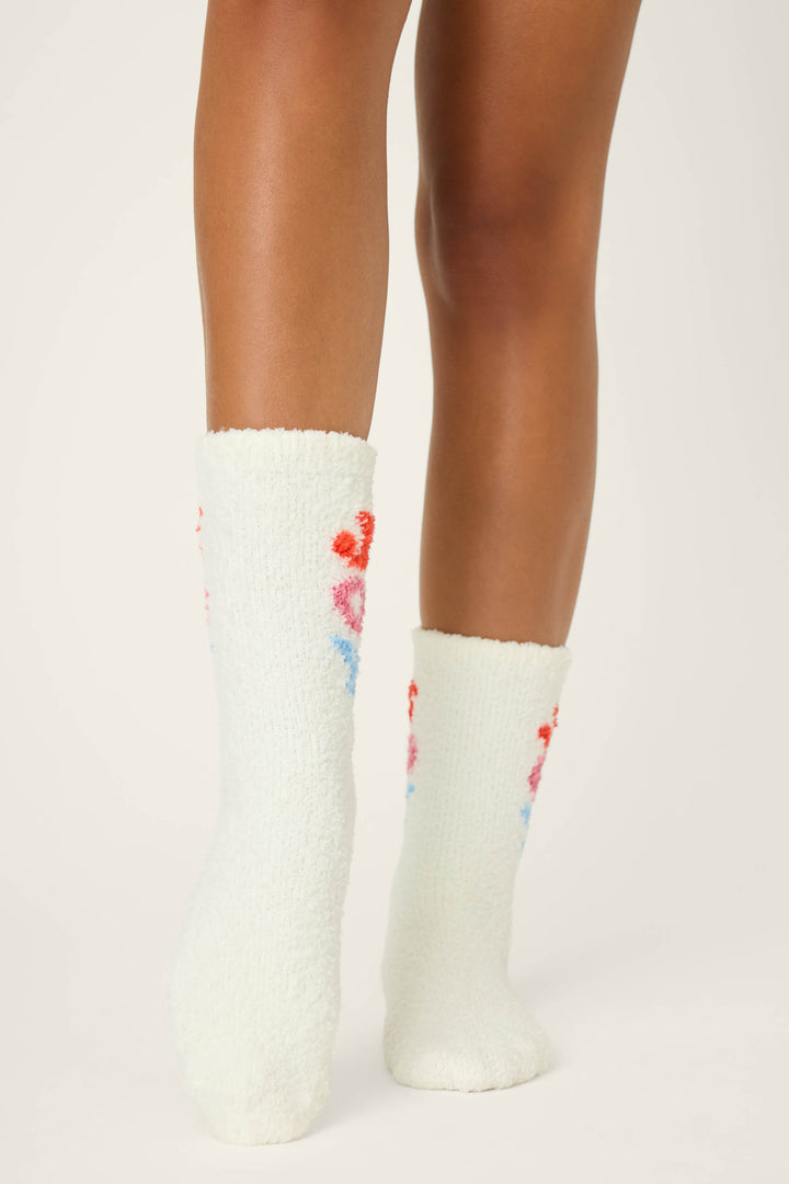 A person with medium brown skin stands with feet together, wearing P.J. Salvage JOY SOCKS SKI JAMMIE CLASSICS—soft white socks with small red and blue floral patterns on the sides—against a plain light background.