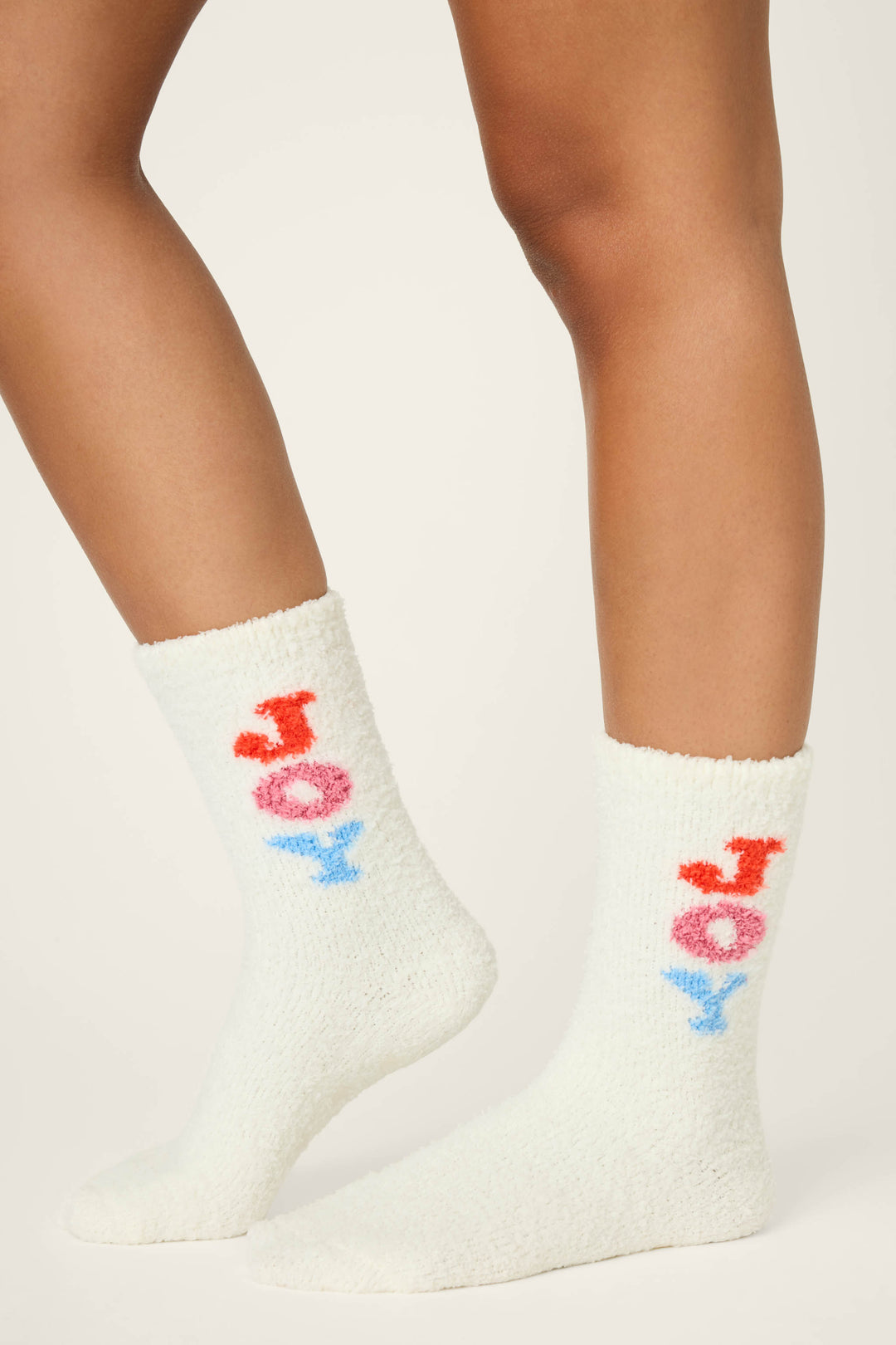 Model showcases the P.J. Salvage JOY SOCKS SKI JAMMIE CLASSICS—fuzzy white socks with "JOY" in red, pink, and blue letters—against a plain light background.