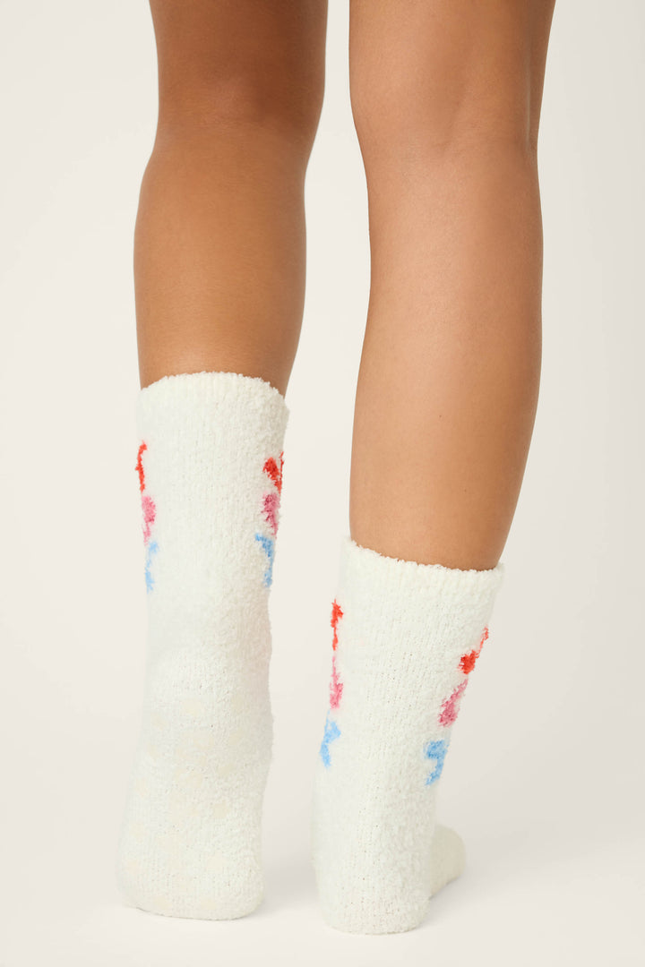 A person stands on a light surface, shown from the calves down, wearing P.J. Salvage JOY SOCKS SKI JAMMIE CLASSICS—soft white fuzzy socks featuring a red, pink, and blue floral pattern on the back.