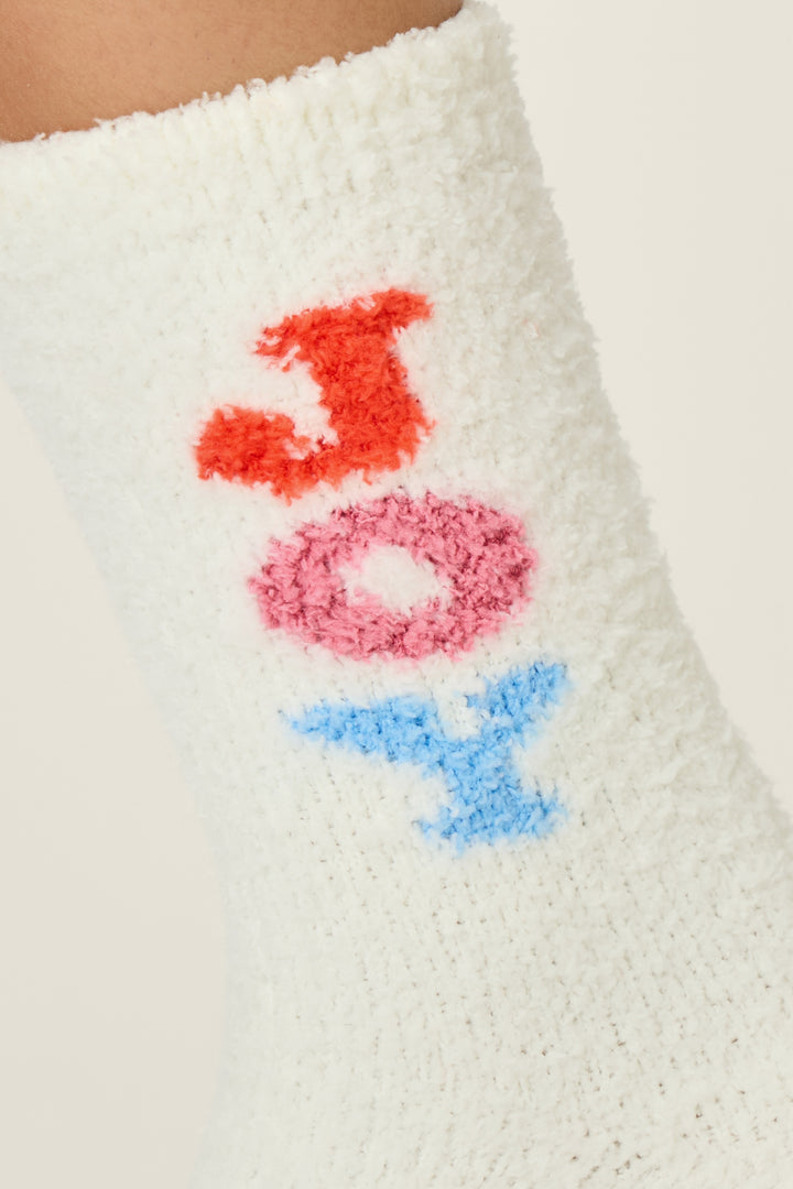 A close-up of P.J. Salvage Ski Jammie Classics Joy Crew Socks with Grippers, featuring white fuzzy fabric and "JOY" written vertically in red, pink, and blue letters.