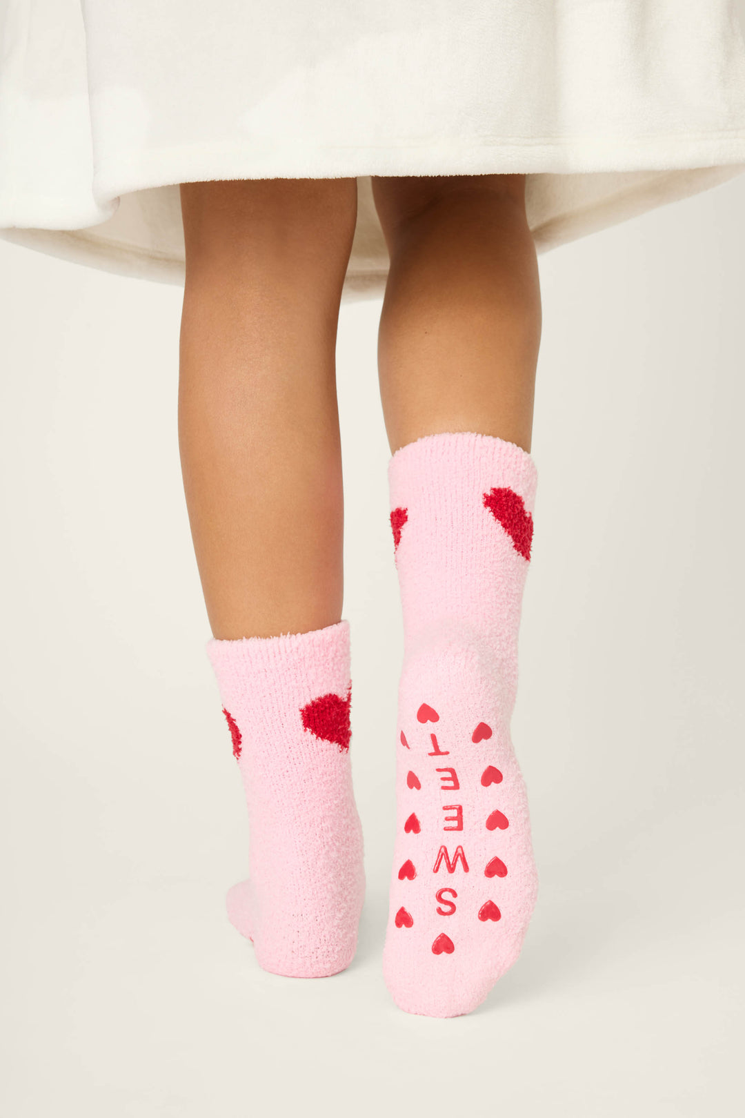 A person stands on tiptoe wearing P.J. Salvage’s Sweeter Than Sugar Heart Print Crew Socks, featuring fuzzy pink fabric, red hearts, gripper bottoms, and “SWEET” in red letters—perfect holiday socks under a white garment.