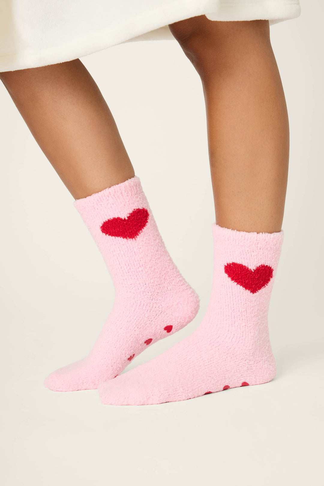 A person in white stands on a light surface, wearing P.J. Salvage Sweeter Than Sugar Heart Print Crew Socks—fuzzy pink socks with red heart details and gripper soles, perfect for cozy holiday comfort.