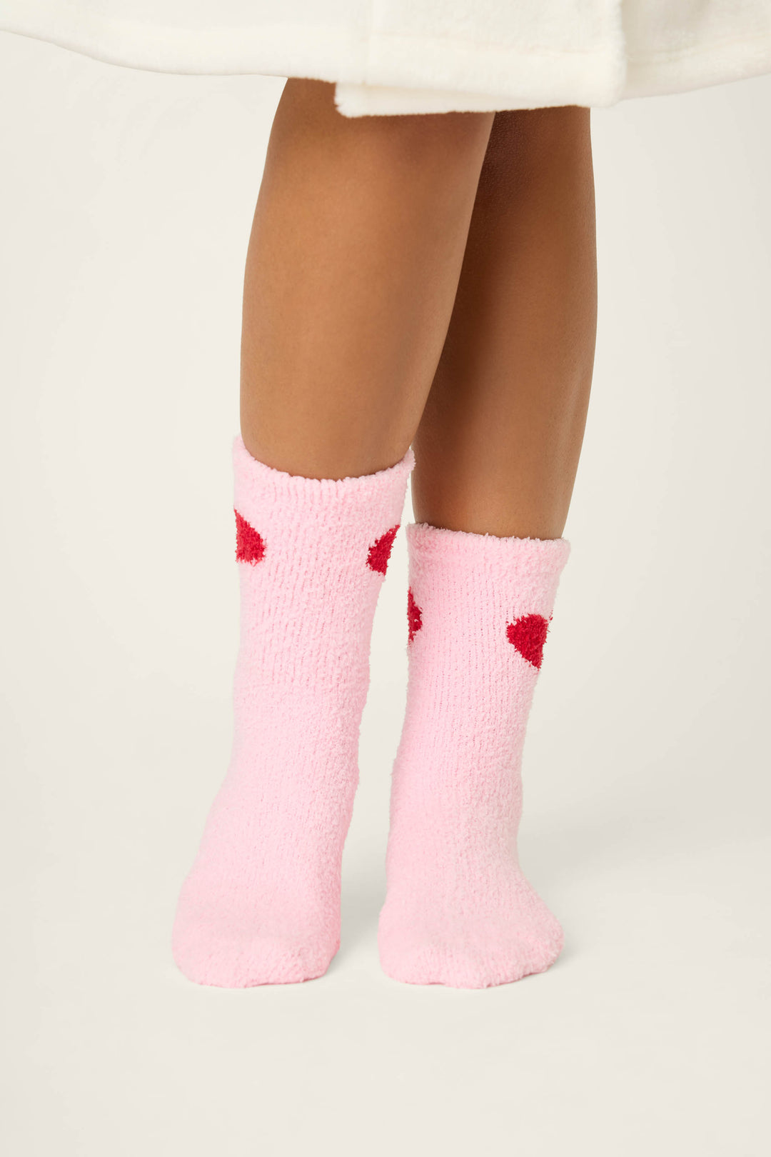A person wearing a white robe stands on a light surface, shown from the knees down, in P.J. Salvage Sweeter Than Sugar Heart Print Crew Socks—fuzzy pink socks with red heart shapes and gripper bottoms for added comfort.