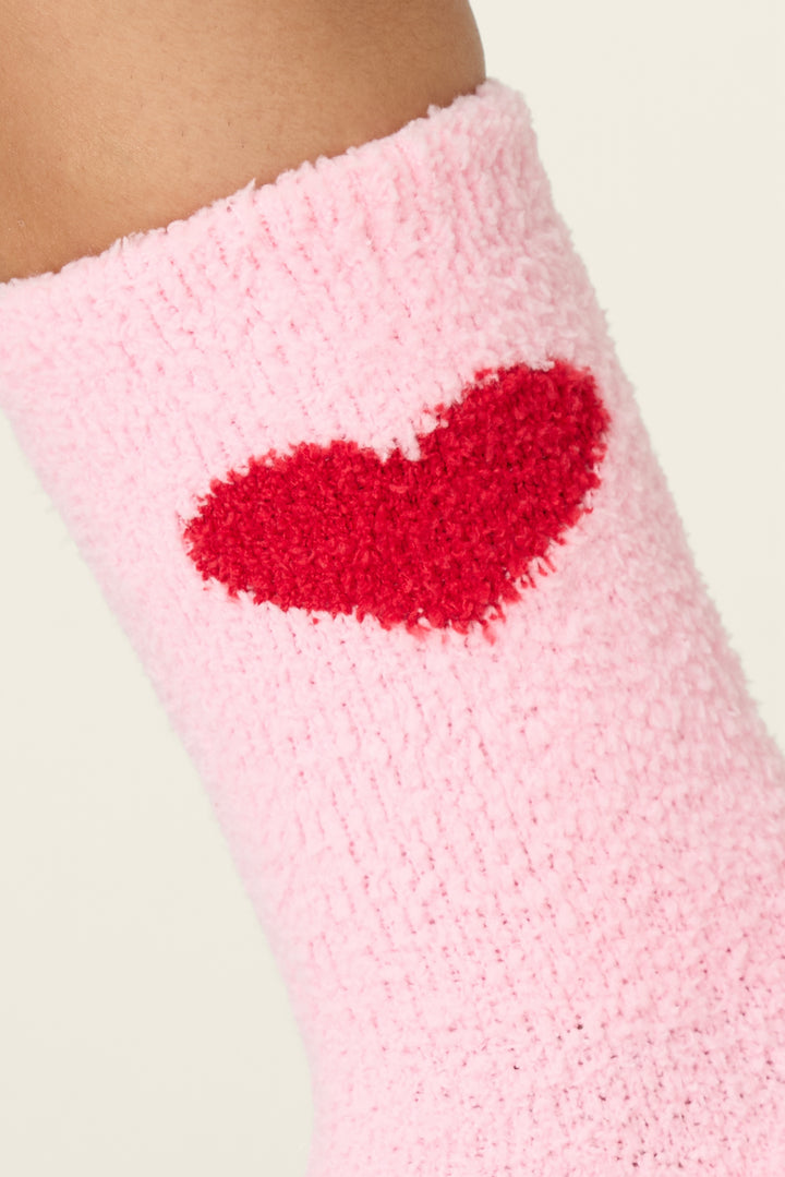 A close-up of P.J. Salvage Sweeter Than Sugar Heart Print Crew Socks, featuring fuzzy pink fabric, gripper bottoms, and a red heart on the side, shown worn on the ankle against a plain light background.