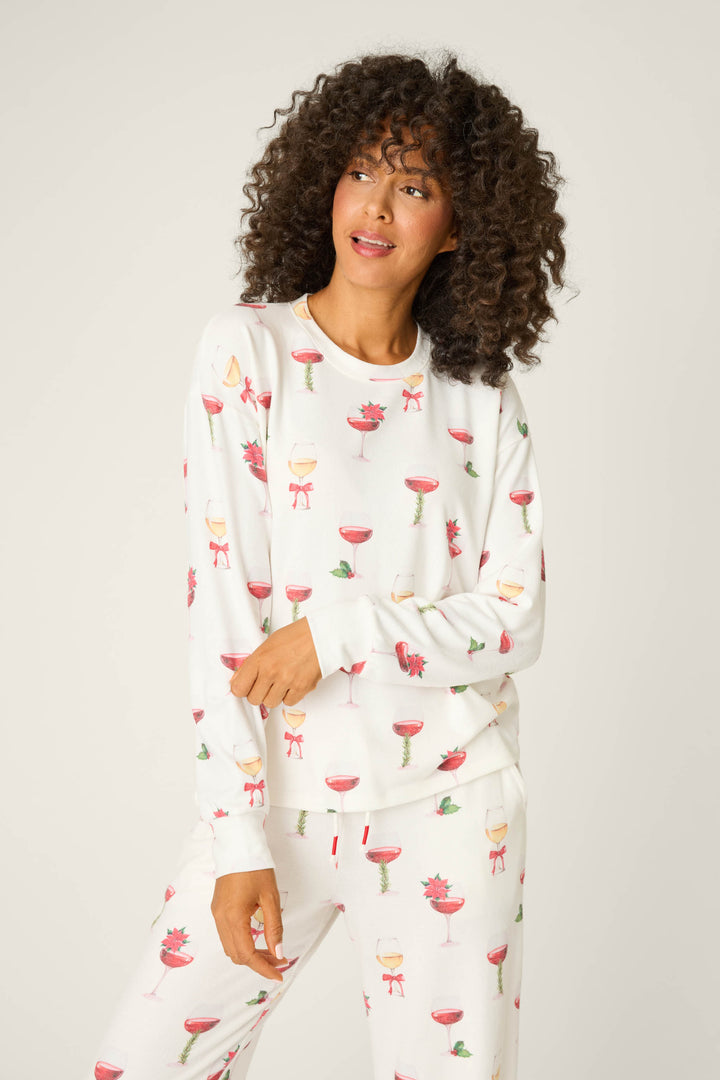 A person with curly hair models the P.J. Salvage Let's Get Toasty Holiday Wine Print Long Sleeve Top, a white pajama set with colorful prints of wine glasses, bows, and flowers, against a plain light background.