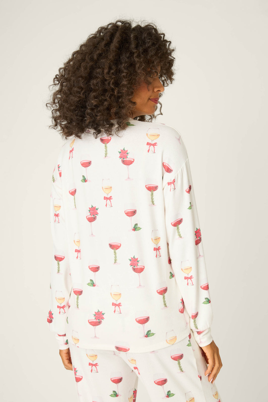 A person with curly hair, turned away from the camera, wears P.J. Salvage's Let's Get Toasty Holiday Wine Print Long Sleeve Top featuring festive drink motifs. The background is plain and light-colored.
