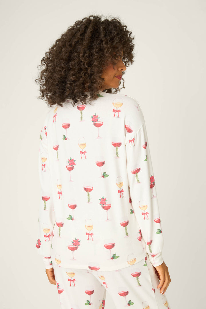 A person with curly hair, turned away from the camera, wears P.J. Salvage's Let's Get Toasty Holiday Wine Print Long Sleeve Top featuring festive drink motifs. The background is plain and light-colored.