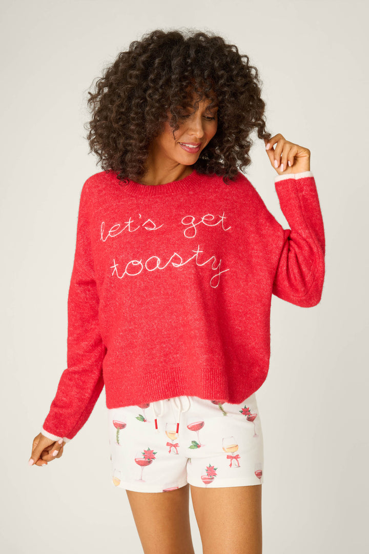 A person with curly hair wears the P.J. Salvage Let's Get Toasty Snuggle Sweater in red and white shorts decorated with holiday drinks and pink accents, standing against a plain light background.