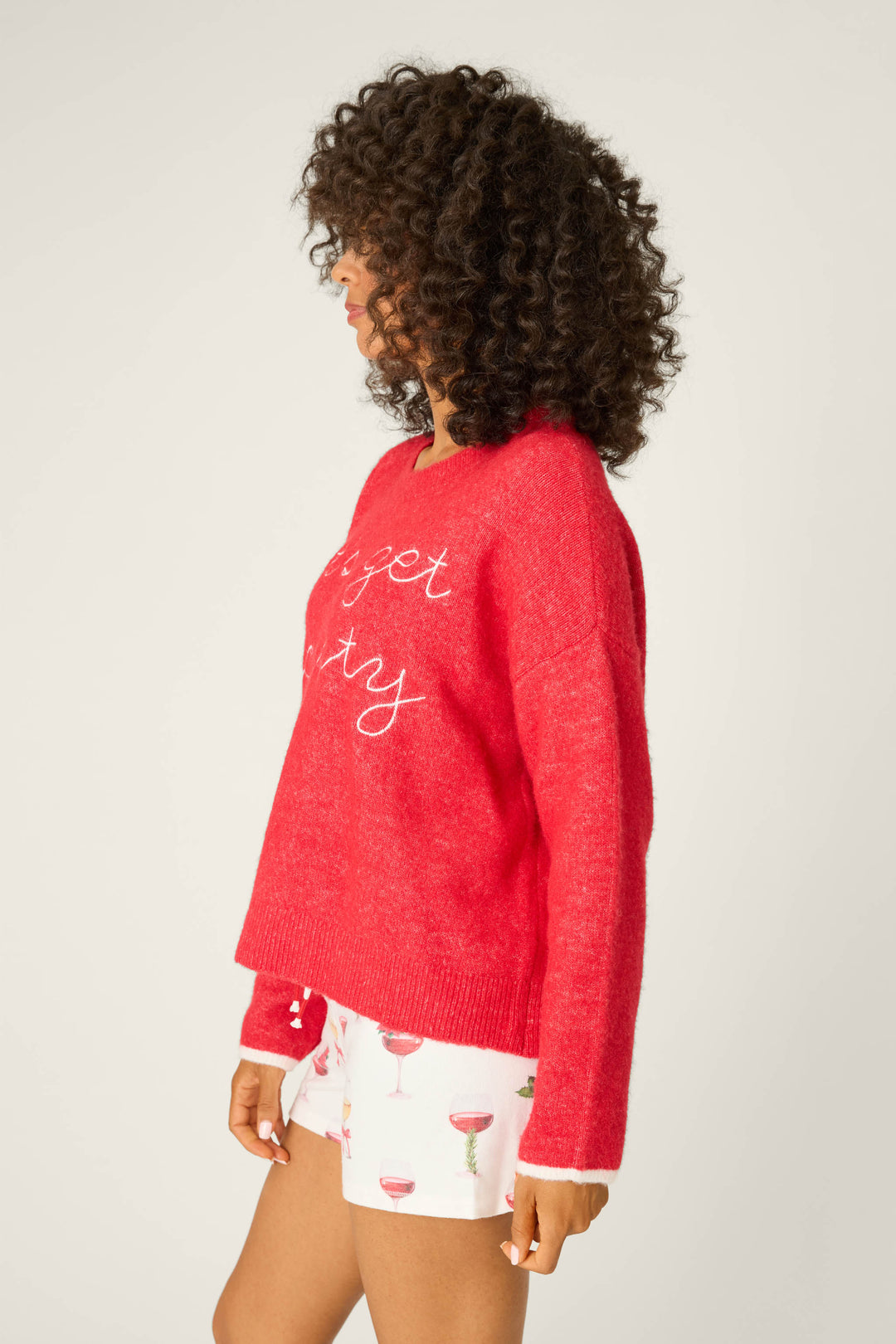 A person with curly hair stands in profile against a plain background, wearing the P.J. Salvage Let's Get Toasty Snuggle Sweater in red with white cursive text, paired with white shorts featuring a cocktail glass pattern.