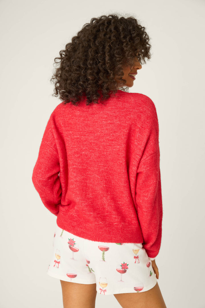 A person with curly hair is shown from the back, wearing the P.J. Salvage Let's Get Toasty Snuggle Sweater in bright red and white cocktail glass-patterned shorts against a plain, light background.