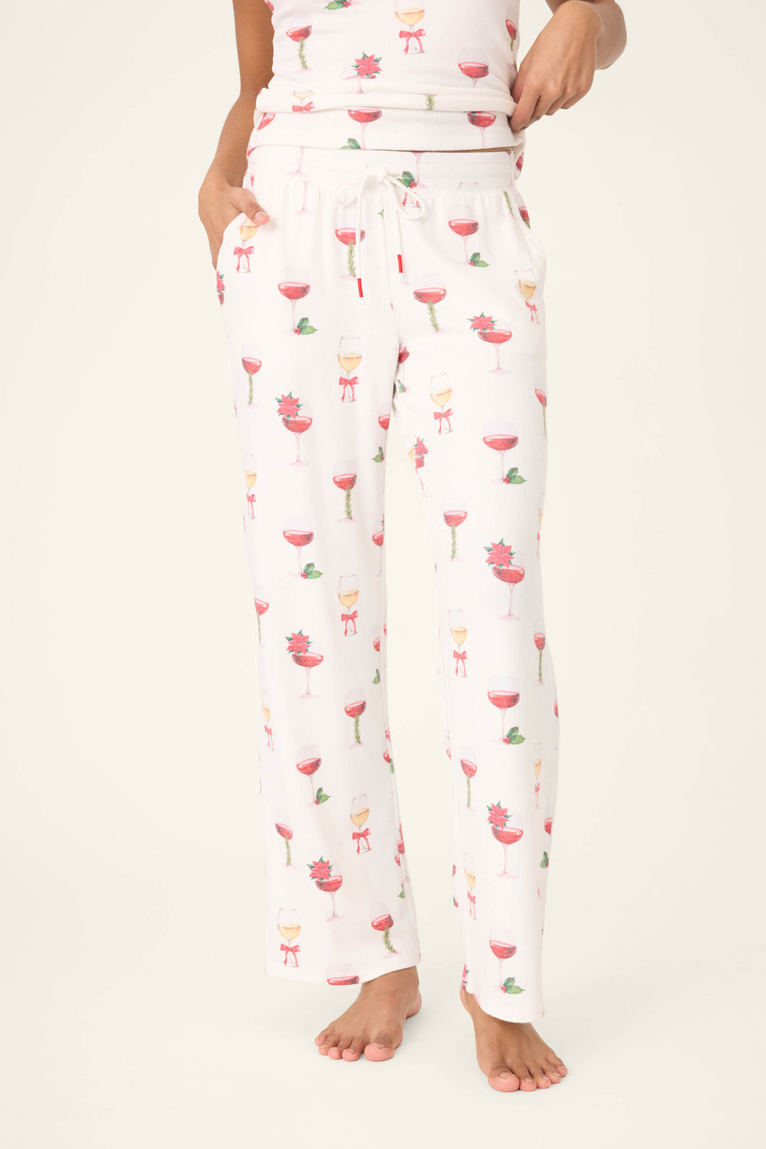 A person is wearing P.J. Salvage Let's Get Toasty Holiday Wine Print Open Leg Pants featuring a drawstring waist, pockets, and colorful wine glass prints, standing barefoot against a plain background.