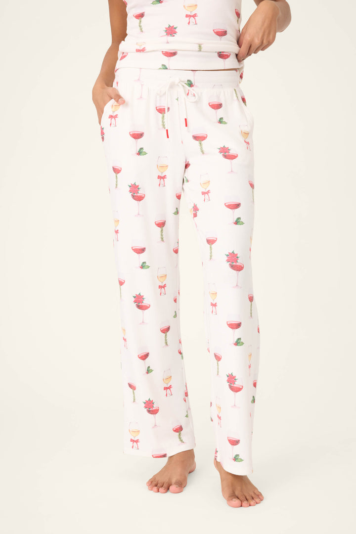 A person is wearing P.J. Salvage Let's Get Toasty Holiday Wine Print Open Leg Pants featuring a drawstring waist, pockets, and colorful wine glass prints, standing barefoot against a plain background.