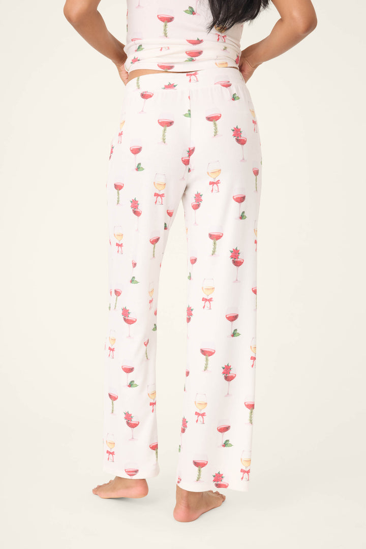 A person stands barefoot, facing away in P.J. Salvage’s Let’s Get Toasty Holiday Wine Print Open Leg Pants, featuring a colorful wine glass and flower pattern. Their hands are on their hips against a plain light background.