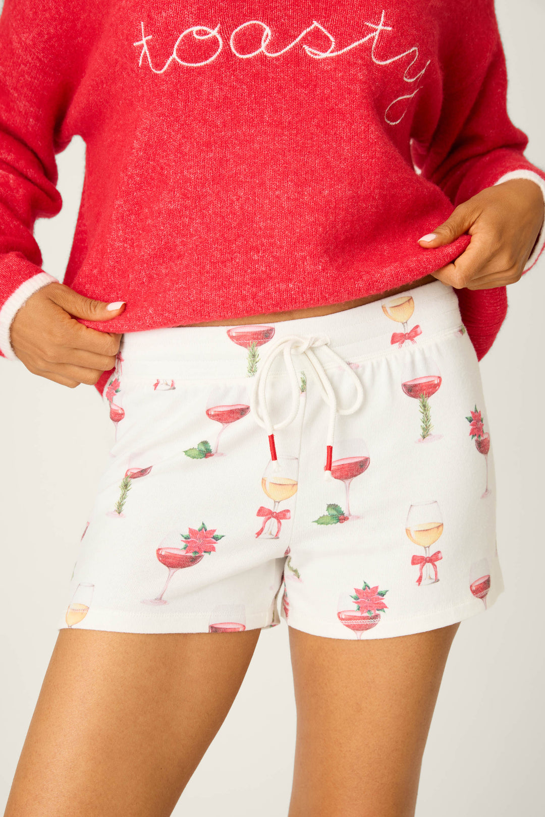 Wearing P.J. Salvage's Let's Get Toasty Holiday Wine Print Shorts, adorned with festive glasses, holly, and bows, a person lounges in style with their hands on the cozy waistband—a perfect holiday look.