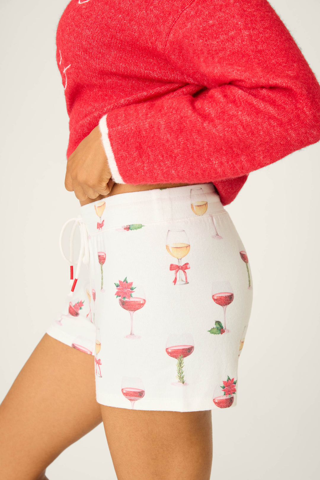 Model wears the P.J. Salvage Let's Get Toasty Holiday Wine Print Shorts—ivory with a festive wine and cocktail print, drawstring waist—styled with a red sweater. Perfect for cozy holiday lounging against a light background.