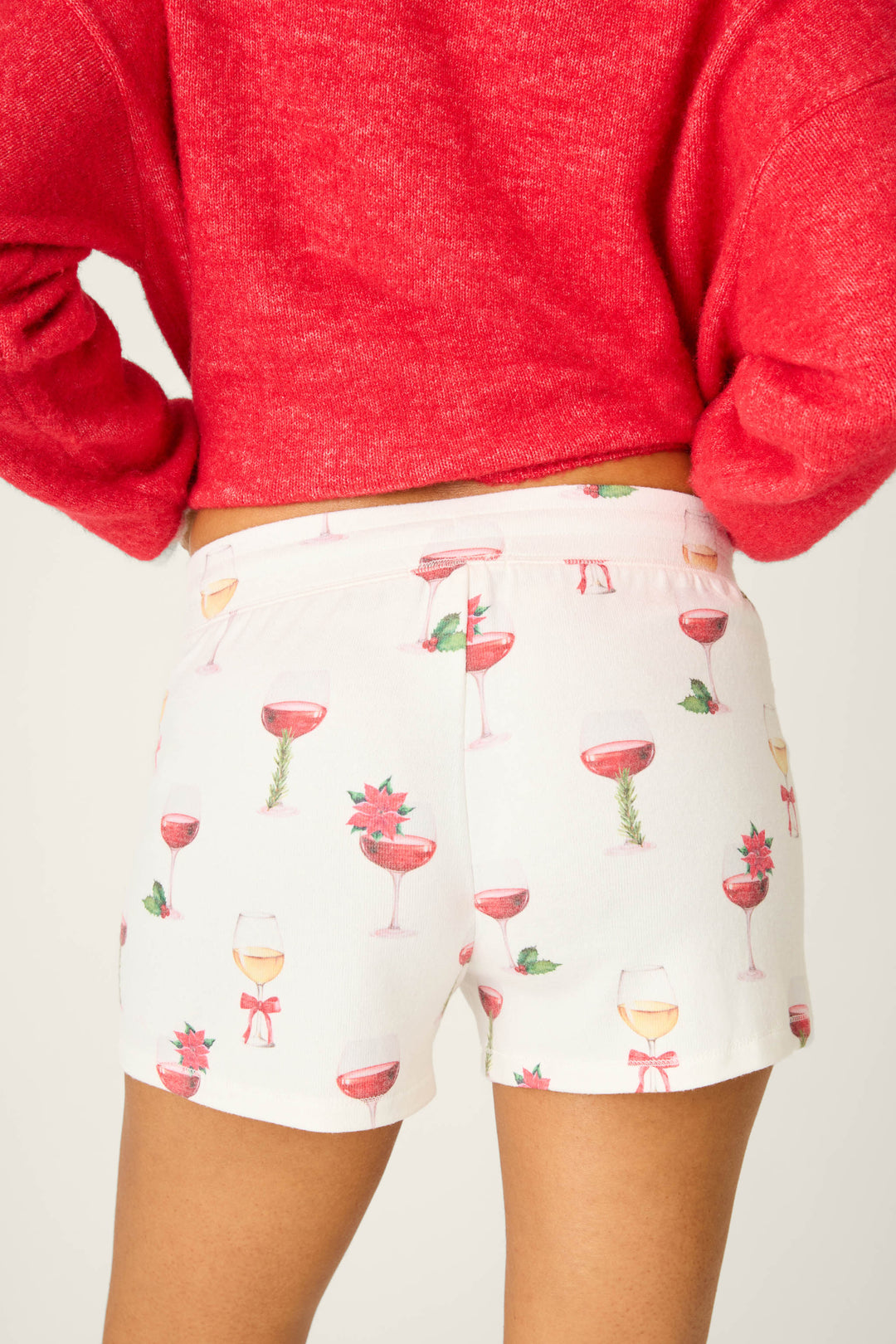 Someone wears P.J. Salvage's Let's Get Toasty Holiday Wine Print Shorts—ivory with festive wine glass prints—paired with a red long-sleeve top, shown from behind and waist down. Perfect for relaxed holiday lounging.