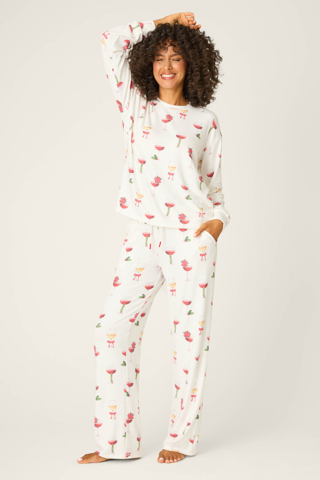A person stands barefoot against a plain background, wearing the P.J. Salvage Let's Get Toasty Holiday Wine Print Pants Set—white pajamas with colorful wine glass prints—one hand in a pocket, the other touching their hair.