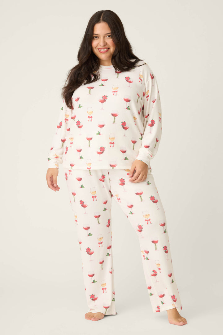 A woman smiles wearing the P.J. Salvage "Let's Get Toasty Holiday Wine Print" plus size pants set, featuring a loose long-sleeve top and matching pants with a festive wine print on a white background. The setting is plain and light-colored.