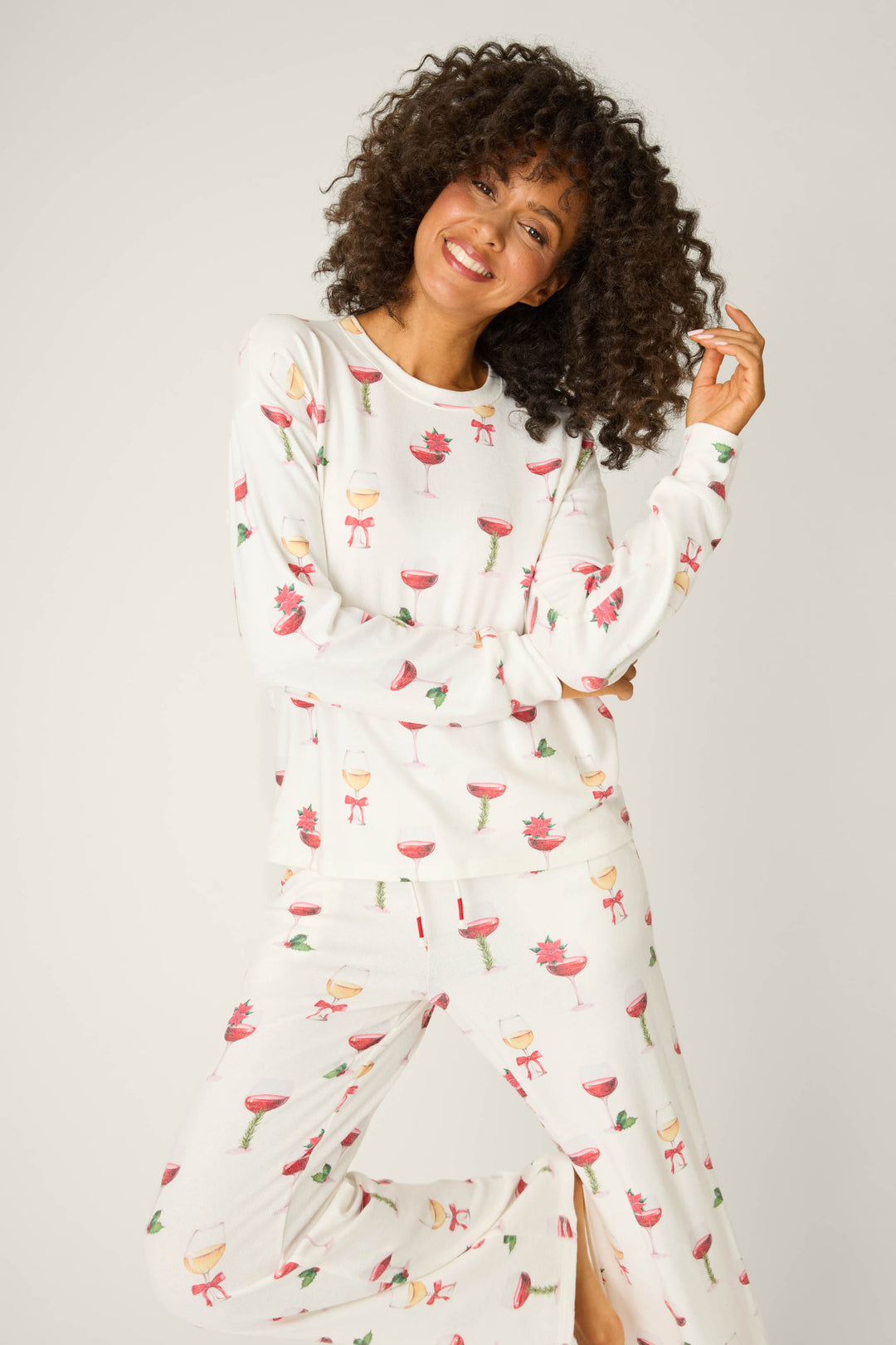 A curly-haired woman smiles in the P.J. Salvage Let's Get Toasty Holiday Wine Print Pants Set, featuring a festive drink pattern, posing against a plain light background.