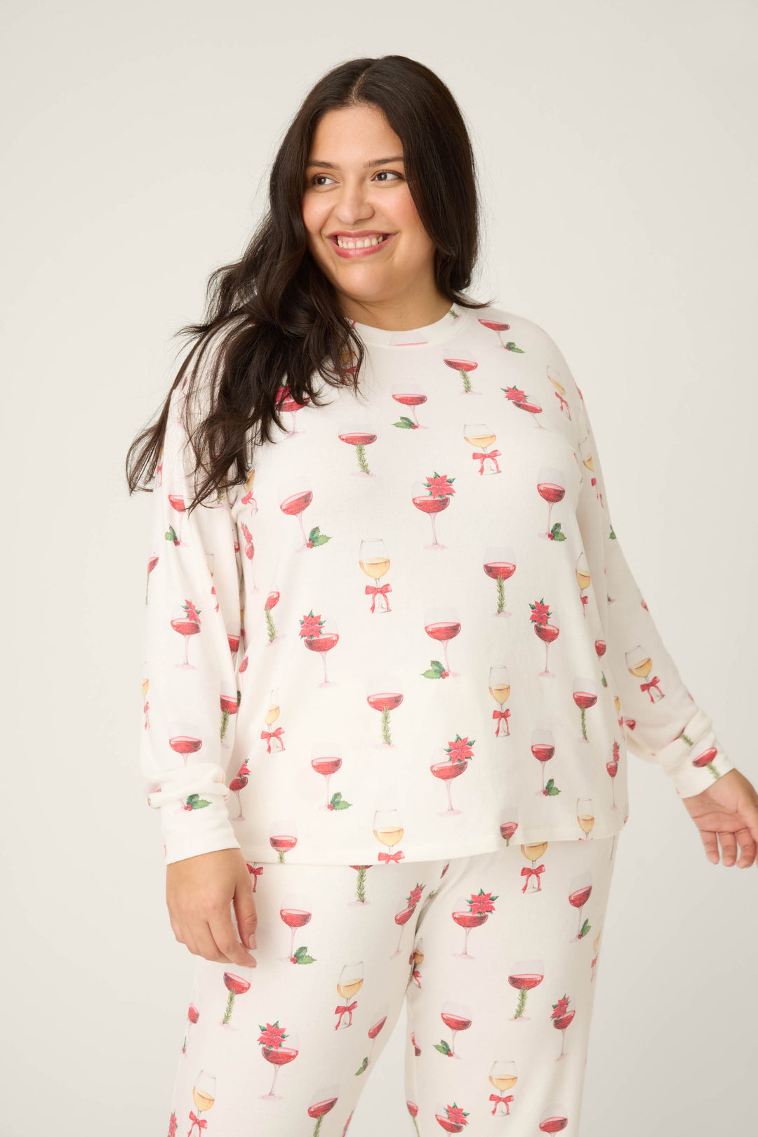 A woman with long dark hair smiles while wearing the P.J. Salvage Let's Get Toasty Holiday Wine Print Plus Size Pants Set, featuring white pajamas with festive red and green wine glasses, poinsettias, and mistletoe prints against a plain background.