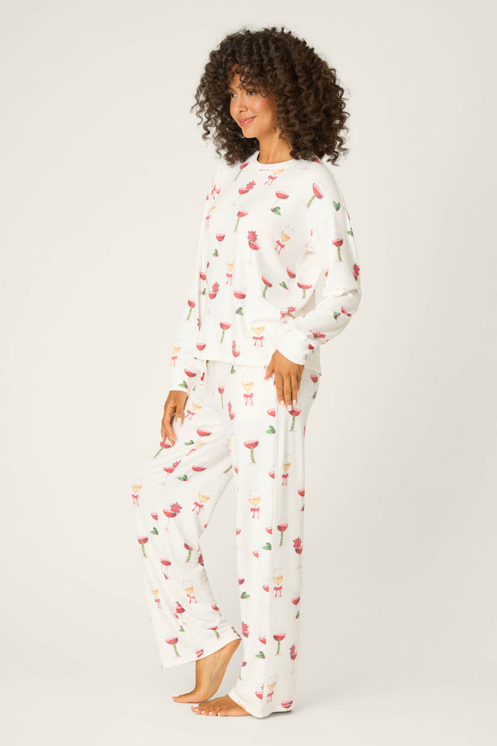 A person stands barefoot on a white background wearing the P.J. Salvage Let's Get Toasty Holiday Wine Print Pants Set—white pajamas with a colorful wine-themed pattern, featuring long sleeves and pants.