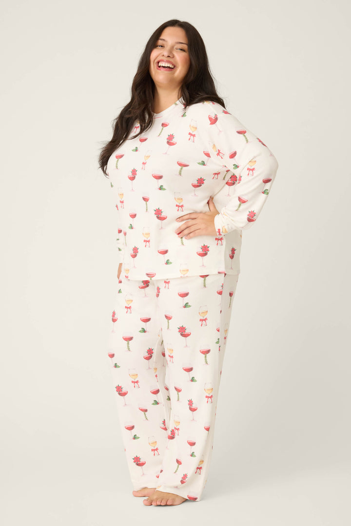 A woman with long dark hair stands barefoot, smiling, in the P.J. Salvage Let's Get Toasty Holiday Wine Print Plus Size Pants Set—white pajamas with colorful drink and people prints—against a light background.