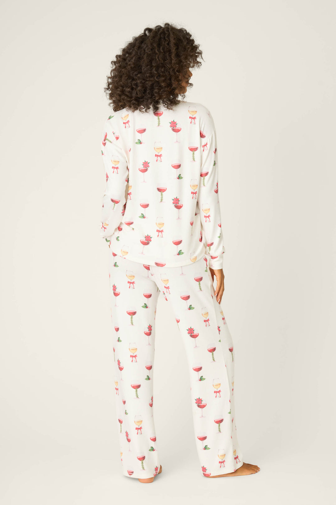 A person with curly hair stands barefoot, facing away, wearing P.J. Salvage's Let's Get Toasty Holiday Wine Print Pants Set—white pajamas with a festive wine-themed print on the long-sleeve top and pants, against a plain light background.
