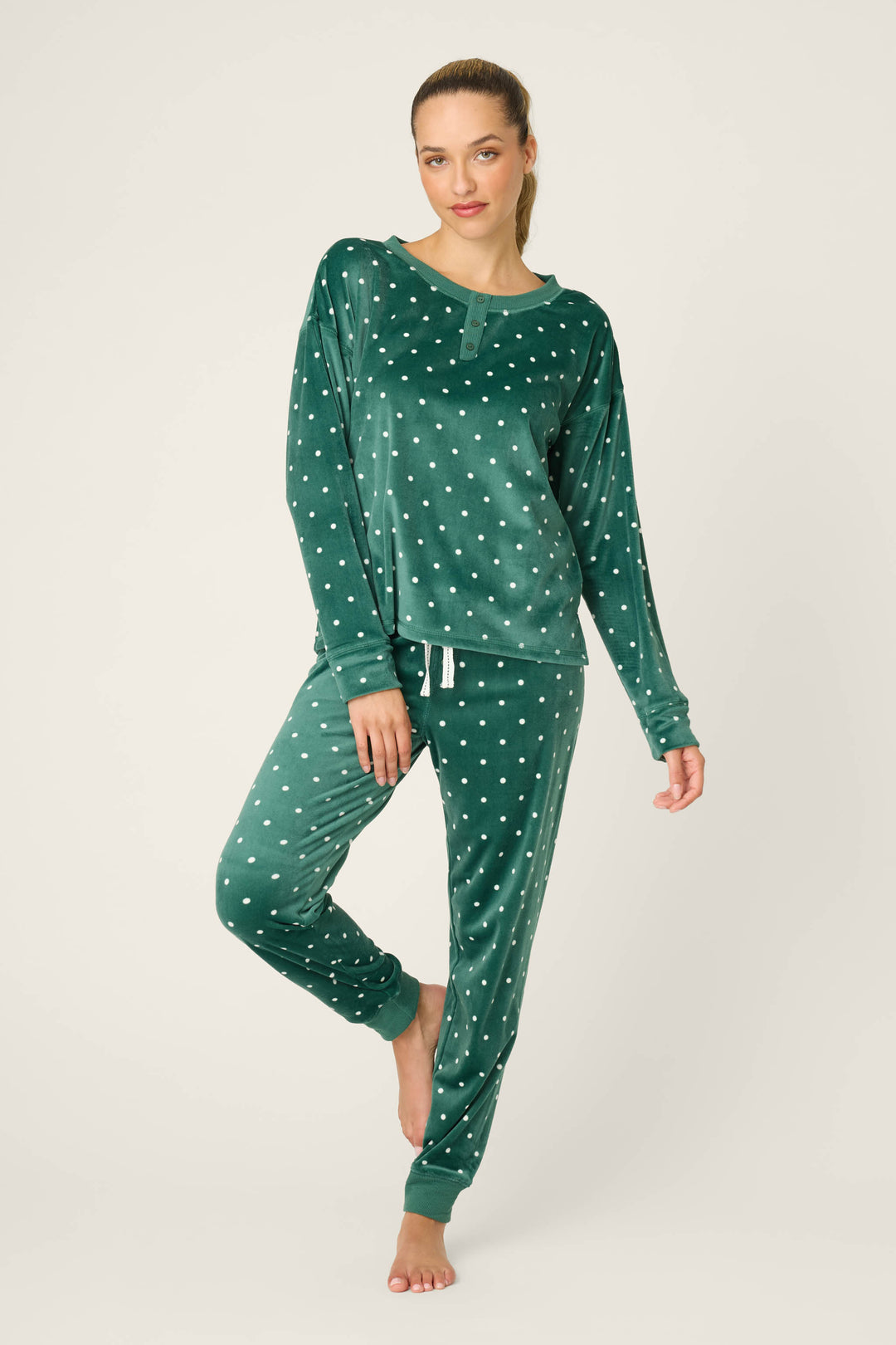A person stands barefoot, posing neutrally in the Let's Get Toasty Dot Print Pants Set by P.J. Salvage—a matching green velour pajama set with white polka dots, featuring a long-sleeve top and drawstring pants, with one leg bent.
