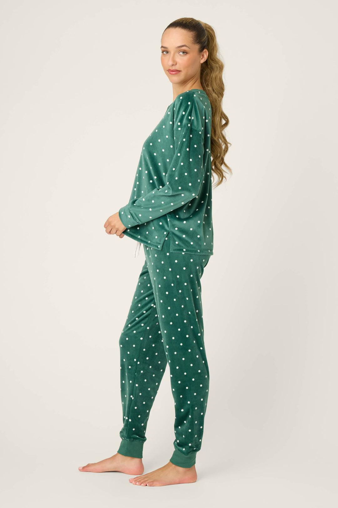 A person stands barefoot in a side pose, wearing the P.J. Salvage Let's Get Toasty Dot Print Pants Set— a matching green long-sleeve top and pants with white polka dots. The background is plain and light-colored.