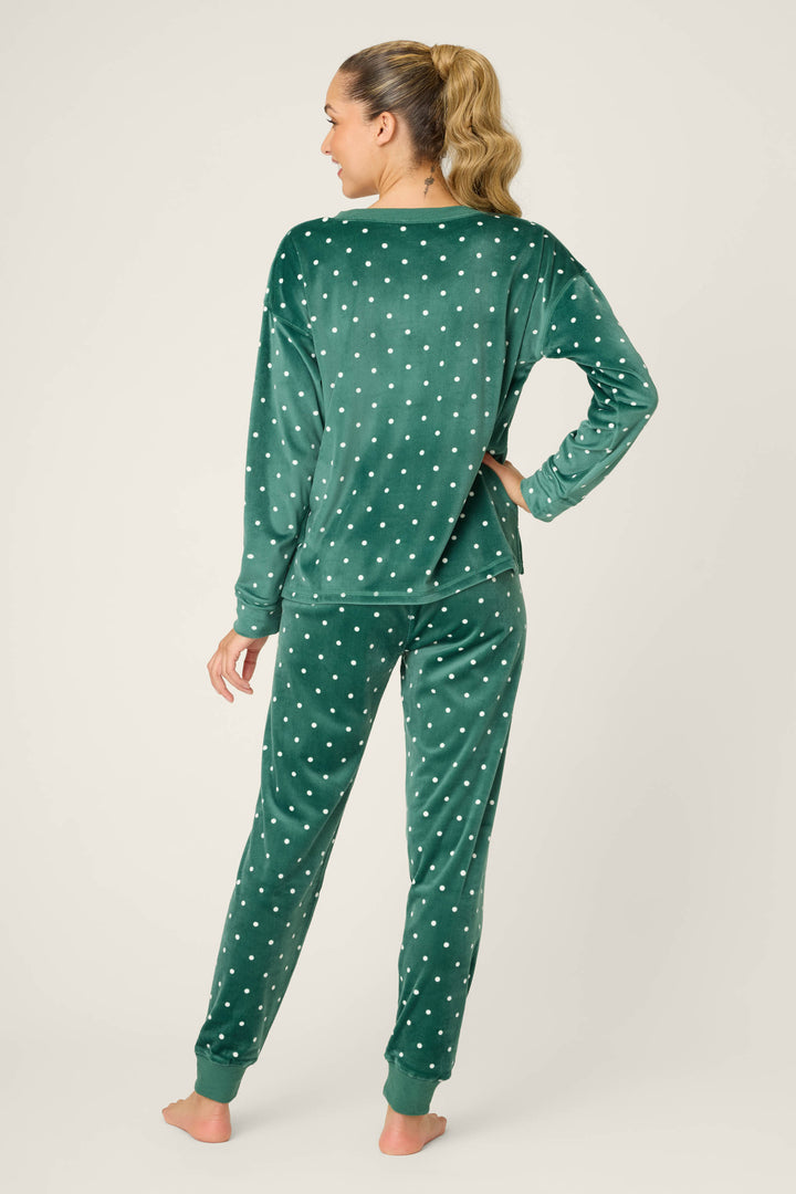 A woman with a ponytail stands barefoot facing away, wearing the P.J. Salvage Let's Get Toasty Dot Print Pants Set—green velour with white polka dots, featuring a long-sleeve top and full-length pants—against a plain, light background.
