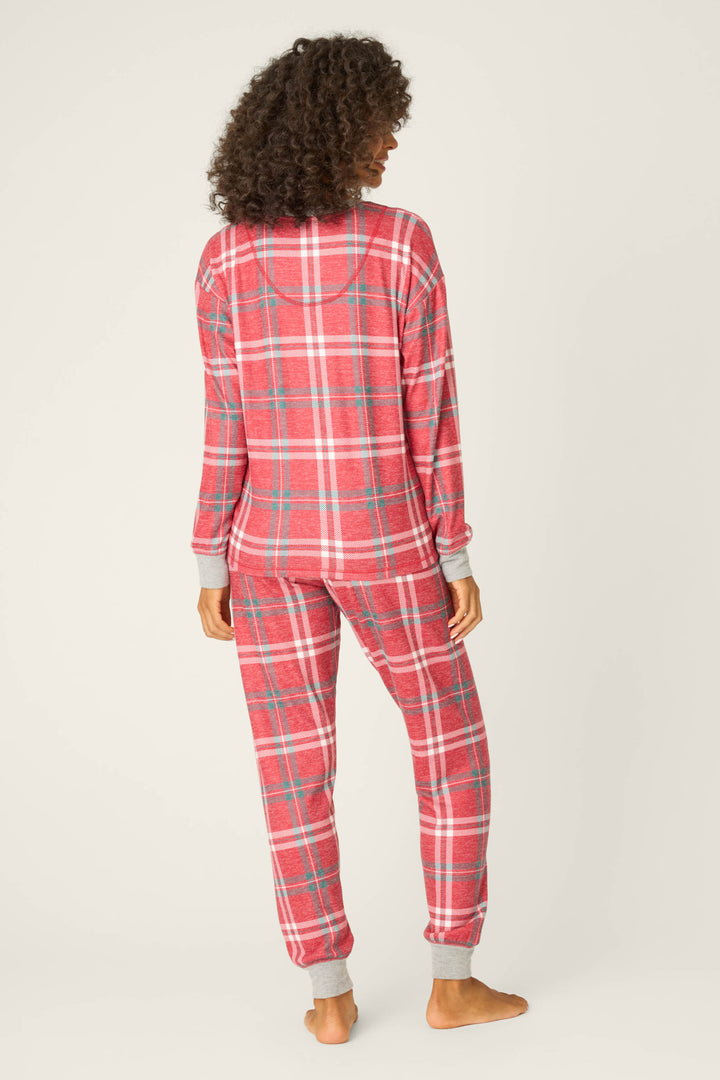 A person with curly hair stands barefoot, facing away, wearing the P.J. Salvage Let's Get Toasty Red Plaid Pants Set, a red and white long-sleeve pajama set with gray cuffs.