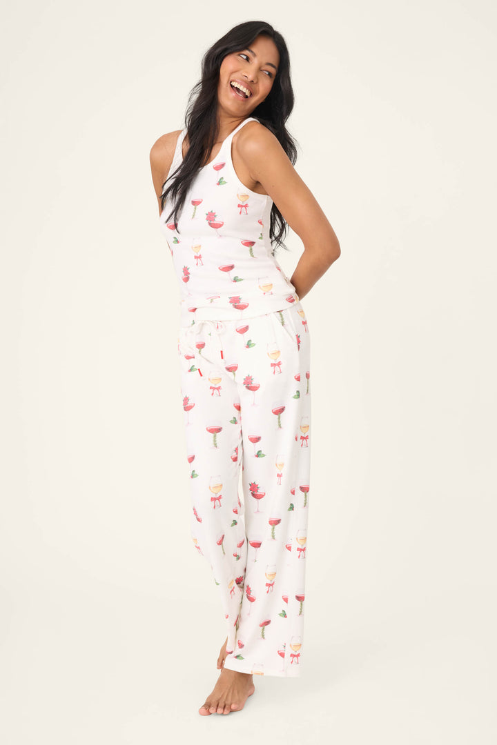 A woman with long dark hair stands barefoot against a plain background, smiling with her arms behind her back. She wears the P.J. Salvage "Let's Get Toasty Holiday Wine Print" tank top in soft jersey fabric.