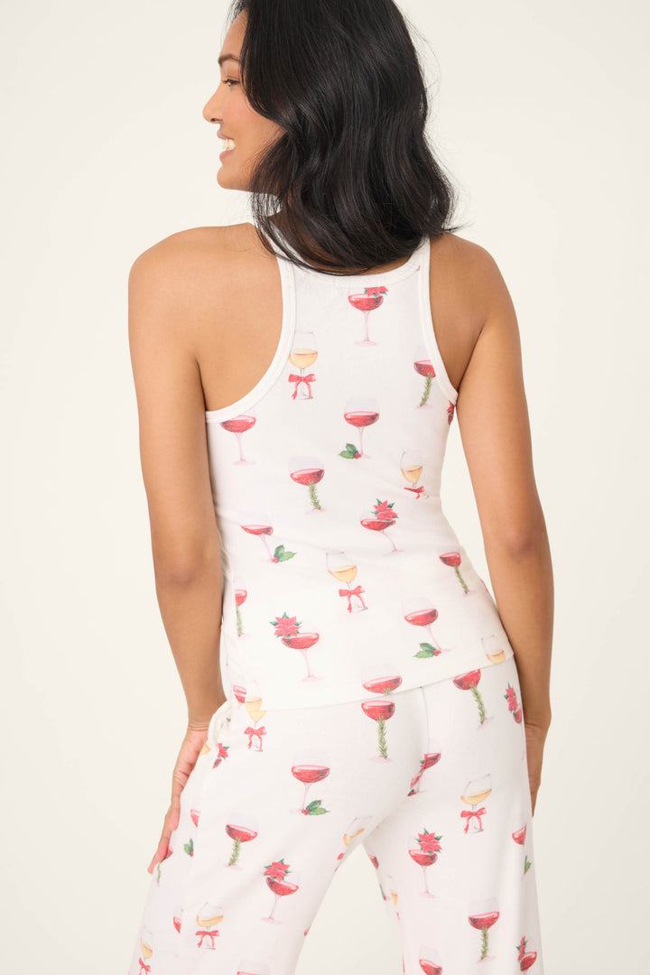 A woman stands against a plain light background, facing away and wearing the P.J. Salvage Let's Get Toasty Holiday Wine Print Tank Top—an ivory racerback style paired with festive pajama pants featuring champagne glass and drink motifs.