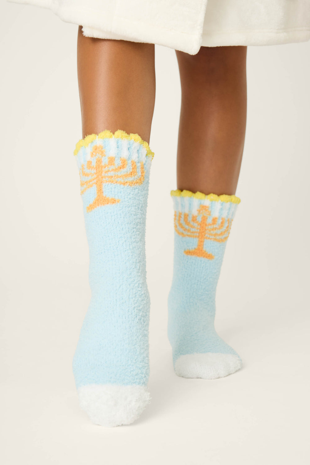A person stands on a light surface wearing P.J. Salvage STARS SOCKS BOYFRIEND BOXER SHORT GIFT SETS: light blue fuzzy socks with orange menorah designs, yellow trim, white toes, partly covered by a white garment.