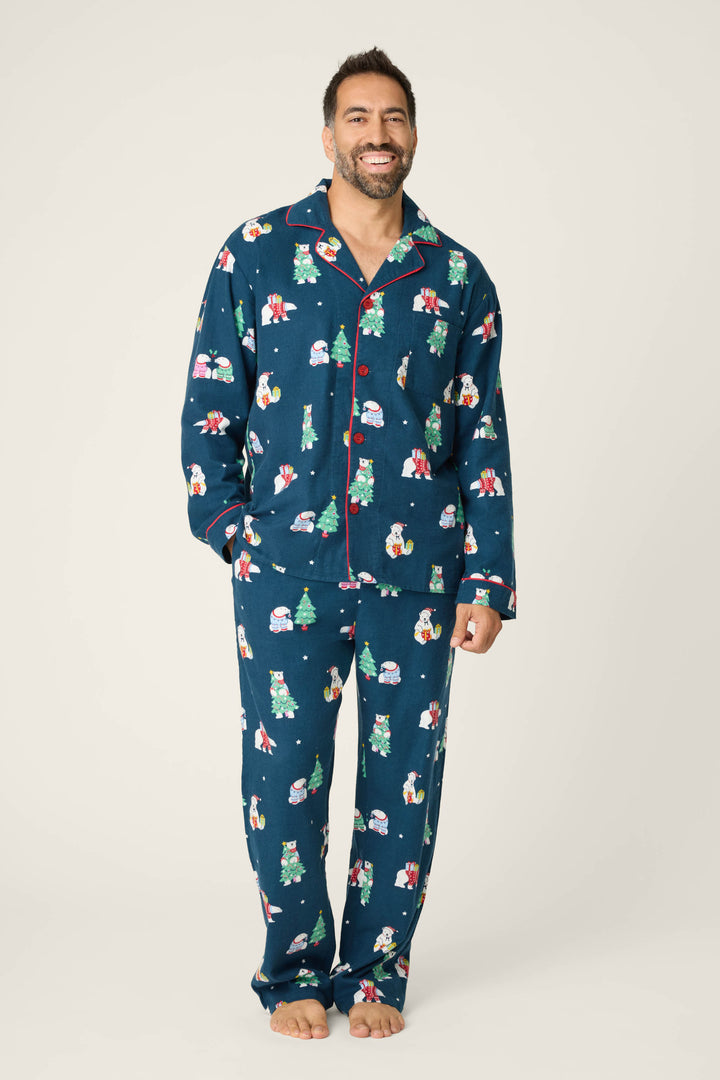 A man smiles barefoot against a plain background, wearing the P.J. Salvage BEARS MENS PJ SET FAM JAM BEARLY AWAKE—navy pajamas decorated with colorful holiday patterns and red edging, one hand by his side.