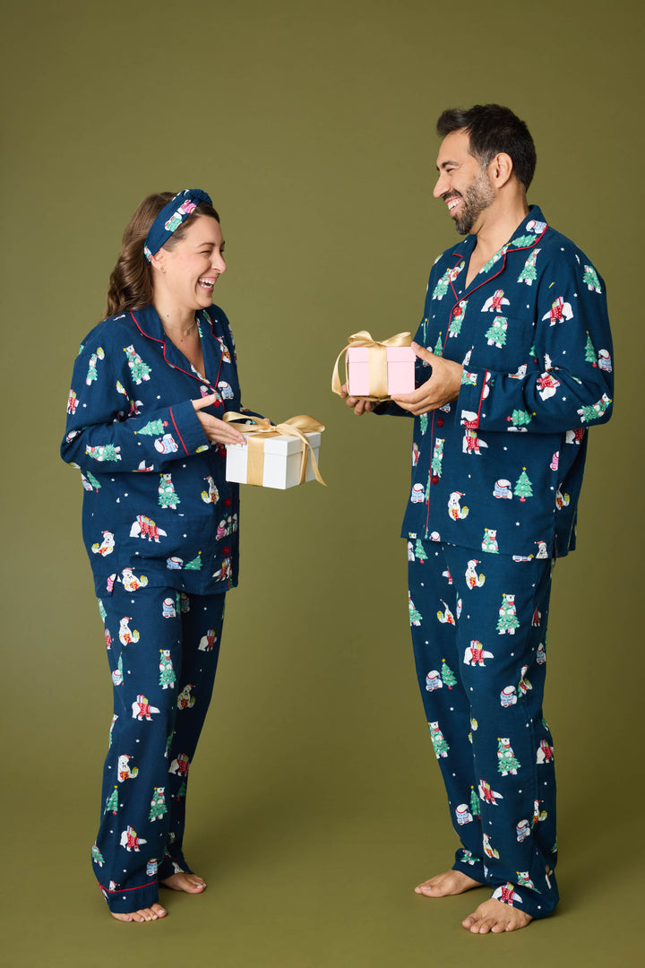 A man and woman wear matching P.J. Salvage BEARS MENS PJ SET FAM JAM BEARLY AWAKE blue holiday pajamas, barefoot on a green background, smiling at each other and holding wrapped gift boxes.