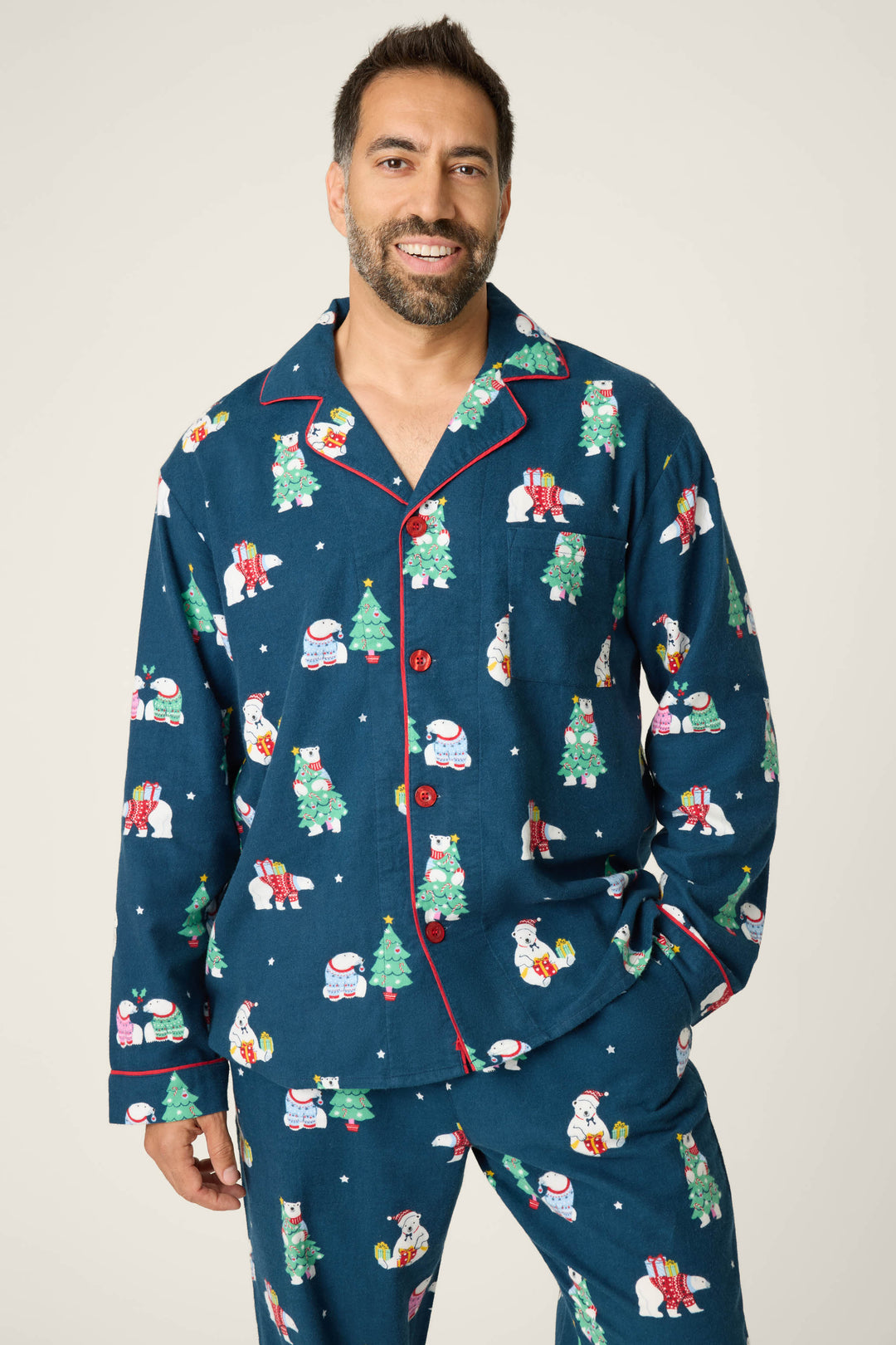 A man with short dark hair and a beard is smiling in the P.J. Salvage BEARS MENS PJ SET FAM JAM BEARLY AWAKE, featuring blue pajamas with festive prints, red piping, and buttons, standing against a plain background.