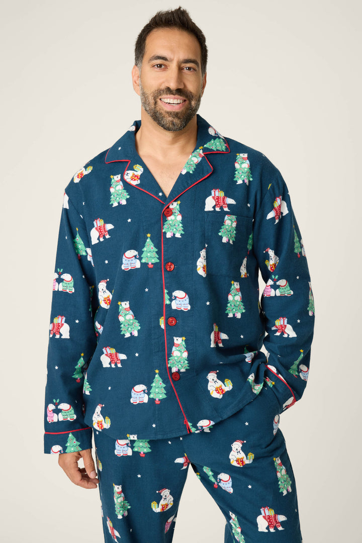 A man with short dark hair and a beard is smiling in the P.J. Salvage BEARS MENS PJ SET FAM JAM BEARLY AWAKE, featuring blue pajamas with festive prints, red piping, and buttons, standing against a plain background.