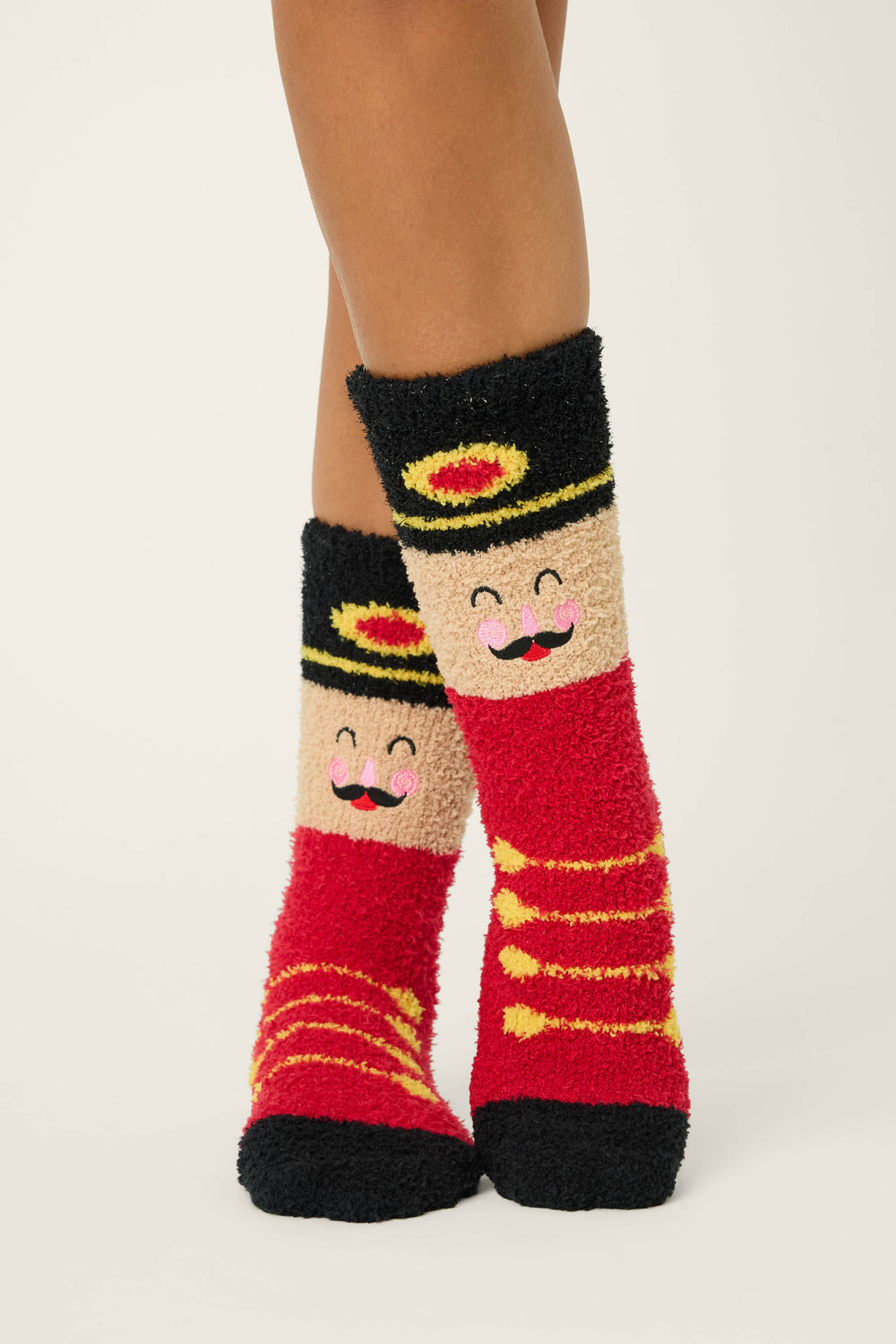 A person wears P.J. Salvage SOLDER SOCKS SKI JAMMIE CLASSICS—fuzzy red socks with toy soldier designs, smiling faces, black hats with yellow and red accents, plus yellow front details. The background is plain and light-colored.