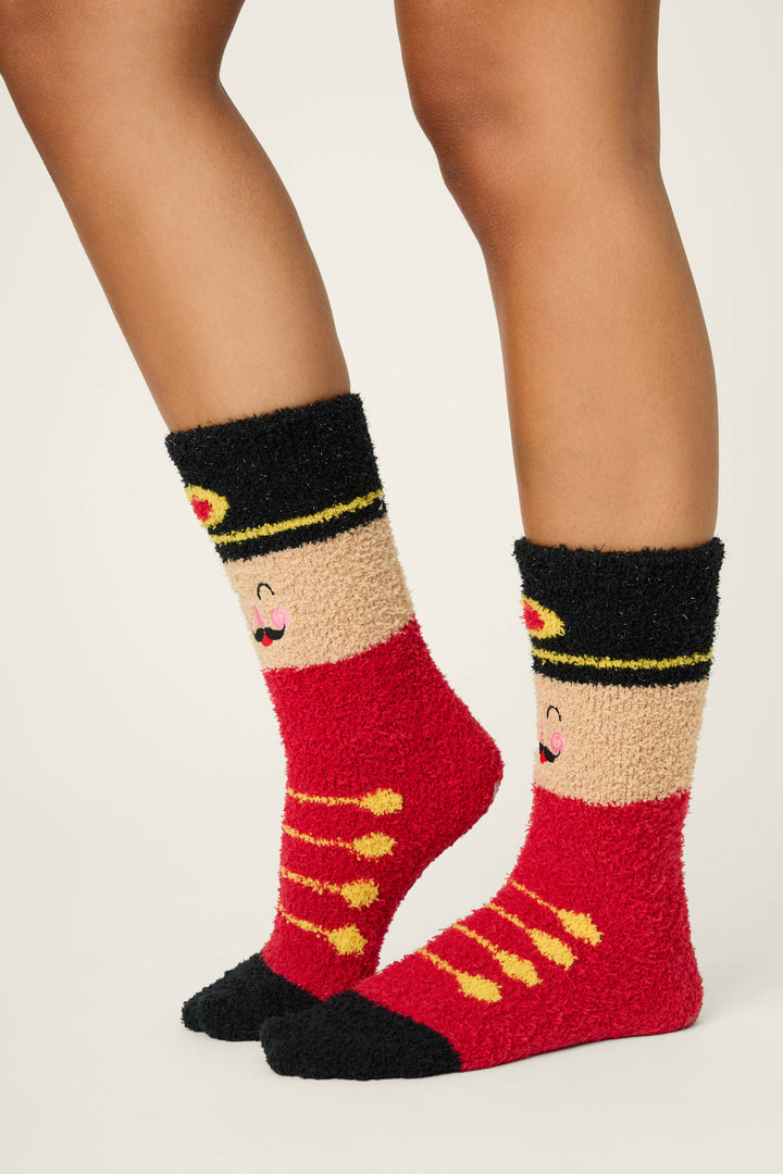 Someone stands on a neutral background wearing P.J. Salvage SOLDER SOCKS SKI JAMMIE CLASSICS—fuzzy red socks with black tops and toes, yellow stripes and buttons, a smiling face near the ankle, and a playful toy soldier design.