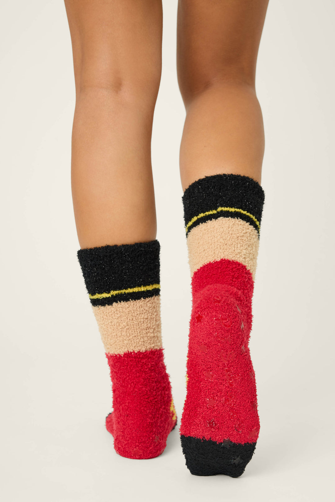 A person stands on a white surface, shown from the knees down, wearing P.J. Salvage SOLDER SOCKS SKI JAMMIE CLASSICS—fuzzy socks with red, black, beige, and yellow stripes.