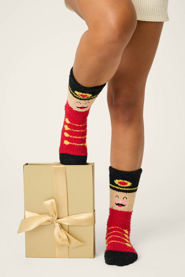 A person in P.J. Salvage SOLDER SOCKS SKI JAMMIE CLASSICS—red, black, and yellow with a smiling toy soldier—rests one foot on a gold gift box tied with satin ribbon, while wearing a white garment.